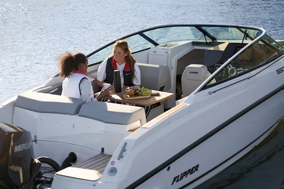 Two women enjoy drinks and snacks on a white boat named "FLIPPER," surrounded by water in a tranquil setting.